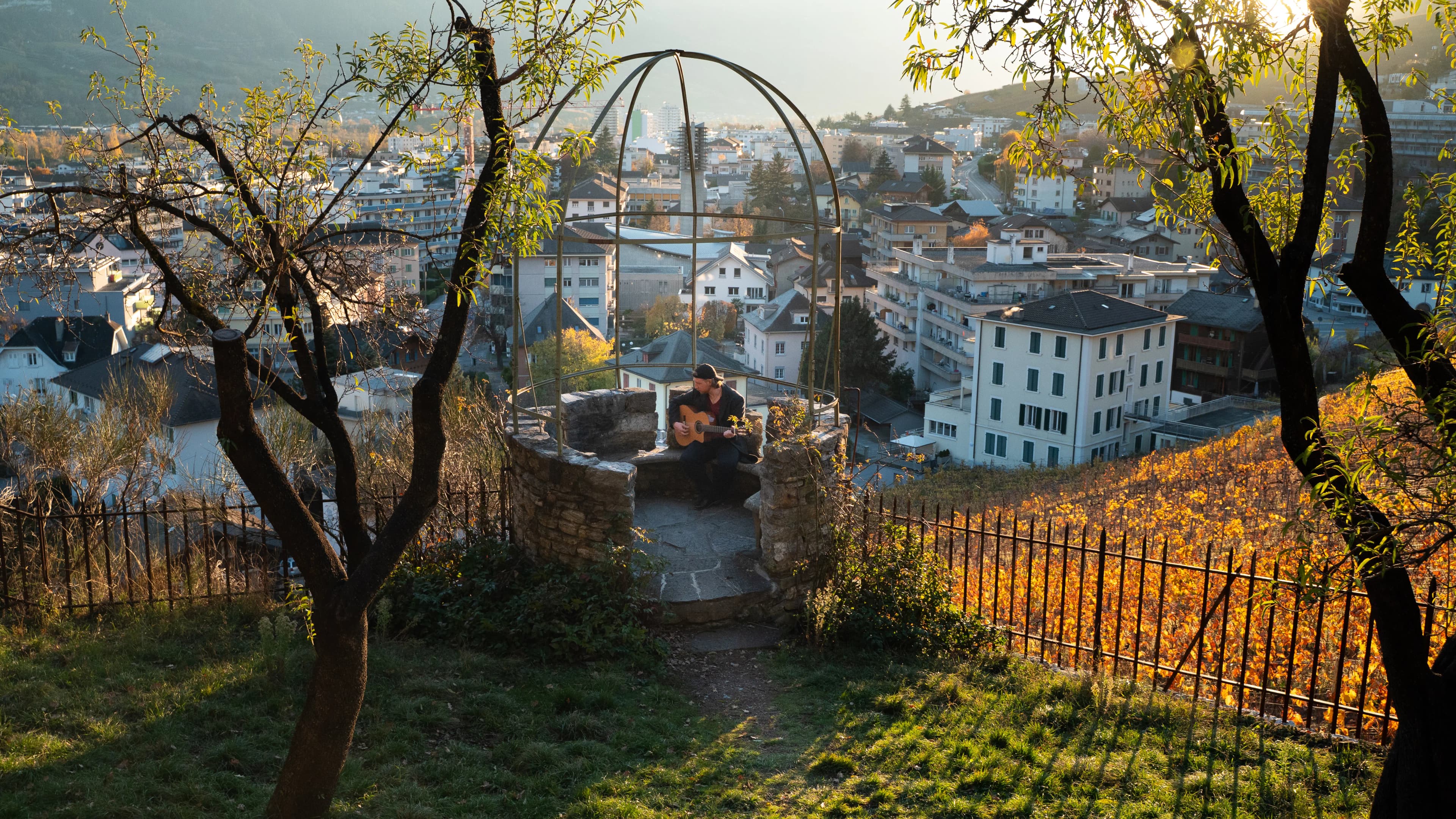 Jakub Bachleda playing guitar in a stone gazebo overlooking a Swiss town and vineyards.