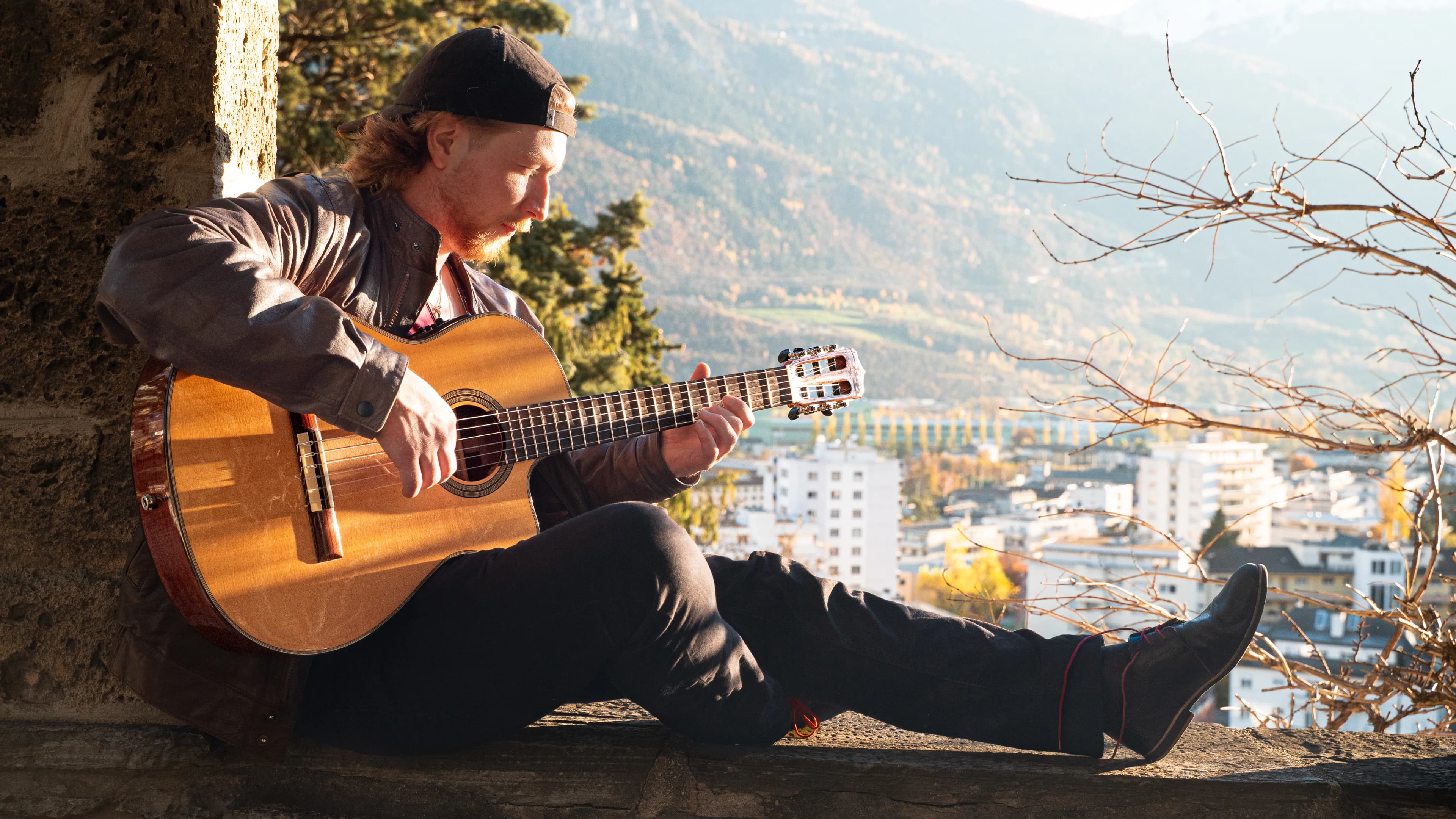 Jakub Bachleda playing guitar above a Swiss town with alpine scenery.